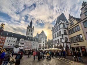 Pretty Places in Germany - This images features Trier's Hauptmarkt, which is a grand plaza with historic Germany buildings located around a beautiful and ornate fountain in the center. There are people walking in the square as well.