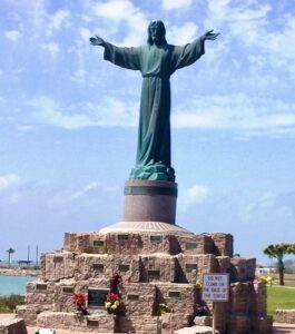 This image features the El Cristo de los Pescadores (Christ of the Fishermen) statue with a bright blue sky in the background.