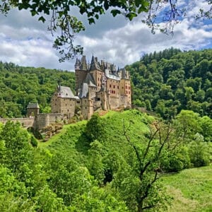 This image features a storybook castle seated on top of a hill surrounded by lush green grass and forest. 