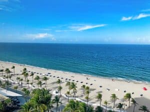 Fort Lauderdale Beach Photo Tour - This image features an aerial view of Fort Lauderdale Beach with Palm Trees swaying in the background. There are beach chairs on the beach and a wide view the pristine blue Atlantic Ocean