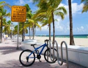 This image features a blue bicycle on the Fort Lauderdale Beach Boardwalk with the curved concrete wall,palm trees and the beach in the background.