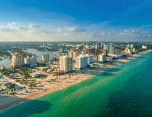 This image features an areial view of the Fort Lauderdale Beach Coastline. The water is emerald green and there are high rise buildings and open beaches on the shore. 