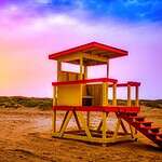 This image features a yellow with red trim life gaurd station on the beach with dunes and a pink and purple sunset in the background.