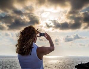 Vacation Photos with Your Phone - This image features a woman taking a photo of a sunset with a digital phone