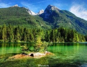 This image features a large twin peak mountain range covered in green trees in the background and an expansive green lake in the foreground reflecting the light of the sun. There is a rock in the lake with trees on top.