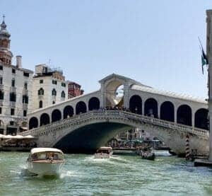 This image features the famous Rialto Bridge in Venice, Italy. There are people standing in the bridge and boats going back and forth under the bridge.