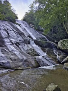 Stone Mountain State Park, North Carolina - Thia image featurea full view of the Stone Mountain Waterfall looking up from where the water pools. There are green trees and large stones and boulders located around the waterfall. 