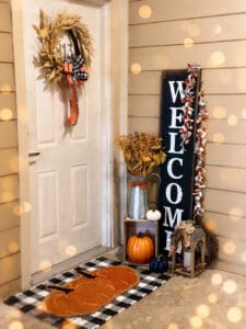 This image features a front porch with a fall wreath on the door and door mat with two pumpkins, and a Welcome sign with a vase with dried flowers, and a few small pumpkins.