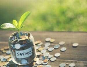Tips for Saving Money for Travel - this image features a glass jar labeled savings that has coins inside and a plant growing in the jar. There are also coins laying on the table that the jar sits on.