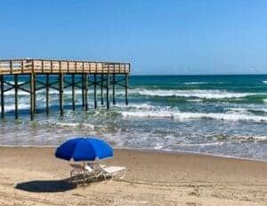 This image features a beach landscape with a wooden pier on the left side and two beach lounge chairs with a blue umbrella.
