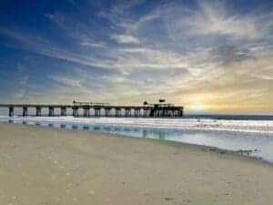 This image features a view of a long pier with a building at the end of the pier with an expansive beach with light waves around at dusk.