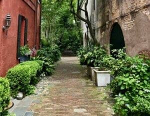 Pretty Places in Charleston - This image is of Philadelphia Alley in Charleston, which shows a rusty red building with black window awnings on the left side and an aged and non updated stone building on the right. There is a cobblestone path between them with trees and green shrubbery all around.