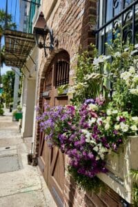 This image features a historic brick building with a brown barn style door that is rounded on top with a lovely flower box of spring flowered on the right of the door.
