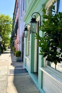 Pretty Places in Charleston - This imagee features a side view of Rainbow Row showcasing the green, purple, and pink facades of the buildings with trees adn black iron glass lights hanging by the doors.
