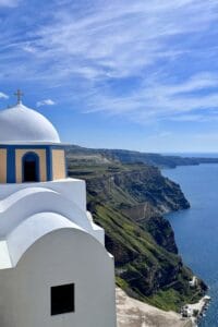 This image features a white domed church on the side of a cliff in Santorini, Greece, overlooking the water below.