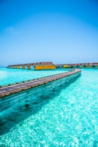 This image features a wooden deck built over bright blue water that leads to cut over the water bungalows.