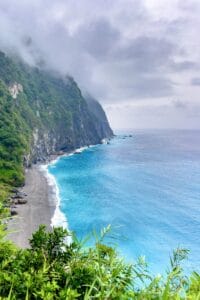 This image features a towering cliff on the left side and a beautiful blue ocean on the right with misty and stormy clouds above.
