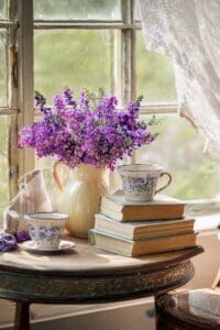 This image features a large pitcher by the window, on a table with purple flowers inside. There is a small teacup to the left and a stack of books and another teacup on the right.