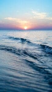 A beach scene with waves flowing onto the shore and the sun setting in the background.