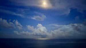 This image features a brightly lit night sky by the moon above. There are wispy clouds at the top of the image by the moon, and fluffy clouds in the middle of the image with the blue water below 