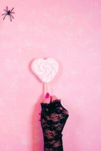 This image features a women’s hand wearing a black lace glove, holding a pink heart lollipop on a pink background with a small black spider in the top left corner. Halloween Aesthetic Wallpapers
