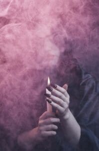 This image features a woman holding a lit candle with smoke billowing around her. She has long black fingernails and the vibe is very witchy - Halloween Aesthetic Wallpapers