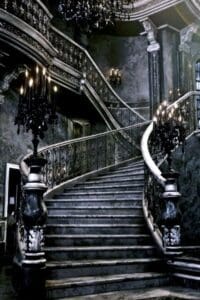 This image is of an vintage staircase in a Victorian style mansion that is dark and dimly lit by candelabras, and is spooky enough to be a witch’s home.