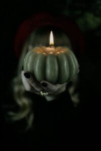 This image features a green pumpkin candle being held by a woman with black nail polish that inspires the idea that she is a witch.