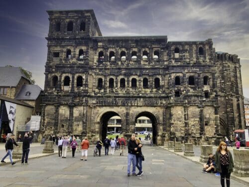 This image features the Porta Nigra in Trier, Germany. It is a large architectural side of a Roman Gate. It features many rounded windows on the upper levels and two lower rounded archways that served as an entry to Trier. There are tourists on the ground beside the gate and they look very small in comparison to the gate.