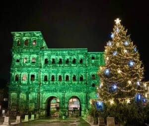 A stunning nighttime photograph of the ancient Roman city gate Porta Nigra in Trier, Germany, lit up in Green with a large decorated Christmas tree on the right hand side of the gate