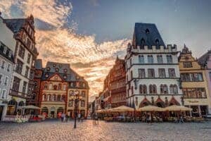 The historic city center square of Trier at sunset. There are German Baroque style buildings with intricate details  cobblestone street with glowing clouds in the sky above.