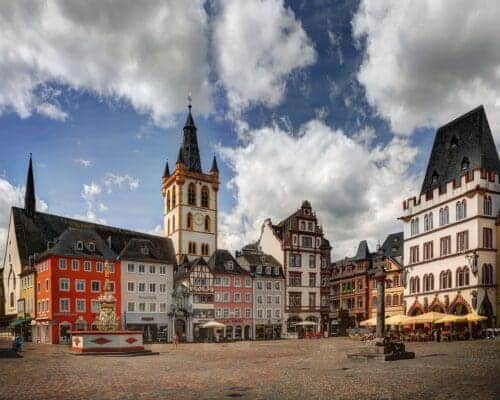 The historic city center square of Trier. There are German Baroque-style buildings with intricate details, a cobblestone street, an intricate medieval fountain, and a blue sky with large clouds.