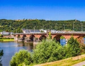 This image features an ancient Roman Bridge in wonderful condition in Trier, Germany. The bridge is made of red brick with arched support structure beneath over the river and a lush green landscape in the background.