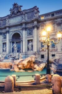 This image features it Trevi Fountian in Rome with a brightly lit lamppost in front.