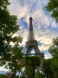 This image features a view of the Eiffel Tower looking up from the ground, with a blue and cloudy sky above and green trees framing the tower.
