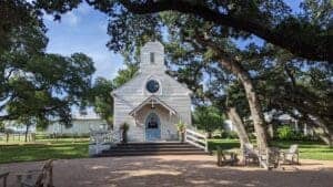 This image features a historic Wooden white church with blue accents under a large oak tree