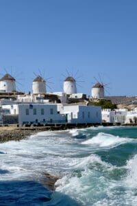 This image features a view of four of the Mykonos Windmills with the blue-green surf rushing onto the beach below them.