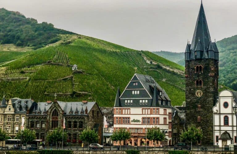One Day in Bernkastel-Kues - This image features a view of th historic town of Bernkastel-Kues, Germany from the water. There are detailed historic buildings lined along the road with deep green vineyards on the hills behind.