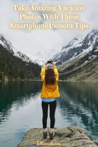 This image is a Pinterest Pin titled, Take Amazing Vacation Photos With These Smartphone Camera Tips. The image features a woman holding her phone high in the air tp capture the snow capped mountain landscape in front of her.