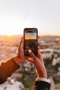 How to take better photos with your phone - Hands holding a smartphone capturing a beautiful sunrise over a scenic landscape.