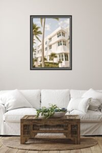 This image features a white Art Deco Building in Fort Lauderdale with Palm Trees in front hanging on the wall in a living room with a white couch.