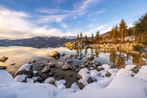 Serene winter landscape at Lake Tahoe with snow-covered rocks and calm water reflecting blue sky.